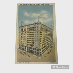 May include: Vintage postcard featuring the John Wanamaker Store in Philadelphia, PA. The multi-story building is depicted with numerous windows, American flags, and the text "100 JOHN WANAMAKER STORE, 13TH AND MARKET STREETS." The postcard has a vintage aesthetic.