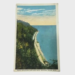 Könnte beinhalten: Vintage Postkarte mit Blick auf den North Boulevard von Arch Rock, Mackinac Island, Michigan. Das Bild zeigt eine Küstenlinie mit Sandstrand, grünen Bäumen und einem blauen See unter bewölktem Himmel. Die Postkarte hat einen weißen Rand.