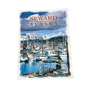 May include: A rectangular postcard featuring a scenic harbor in Seward, Alaska. The image displays numerous white boats docked in blue water, with snow-capped mountains in the background under a cloudy sky. The text SEWARD ALASKA is printed at the top.