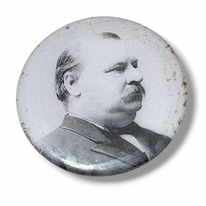 May include: A circular pinback button featuring a black and white portrait of a man with a mustache in profile. The button has a glossy finish with visible rust-colored spots along the right edge and the text Graphics and More on the left.