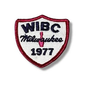 May include: A shield-shaped embroidered patch with a white background and a dark red border. The text WIBC is in dark blue, followed by the word Milwaukee in dark blue script, a pink bowling pin icon, and the year 1977 in dark blue text.