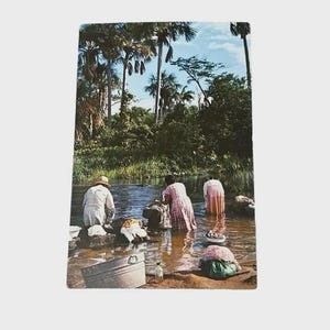 Op de afbeelding: Vintage ansichtkaart met mensen die kleren wassen in een rivier. De scène toont verschillende individuen in het water, omringd door weelderig groen en hoge palmbomen. Een metalen bak en andere items zijn zichtbaar.