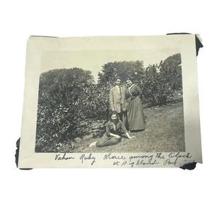 May include: A vintage black and white photograph showing three individuals in a garden setting. One person sits on the grass while two others stand behind. Handwritten text at the bottom reads: Vahon Ruby Marie among the lilacs at Highland Park.