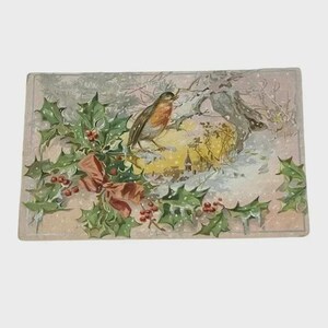 Puede incluir: Tarjeta de Navidad rectangular vintage con un petirrojo posado en una rama con acebo y bayas. El fondo representa una escena invernal nevada con un edificio y un lazo rojo.