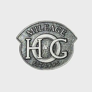 May include: A silver-colored metal pin with the words "MILEAGE HOG" and "EST 1995" in a circular design. The pin has a textured background and a shield-like shape, with the letters "HOG" prominently displayed.