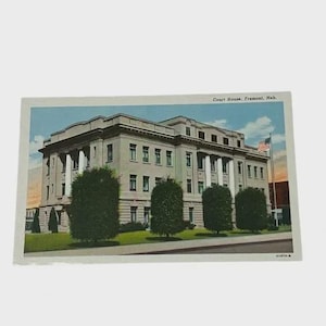 May include: Vintage postcard featuring the Court House in Fremont, Nebraska. The building is a large, light-colored structure with columns and multiple windows. The postcard has a blue sky background and a green lawn in front of the building.