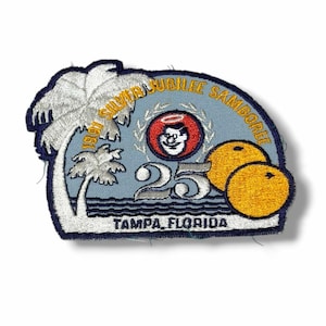 May include: A semi-circular embroidered patch featuring a light blue background with white palm trees on the left, two orange citrus fruits on the right, and the text 1991 SILVER JUBILEE SAMBOREE, 25, and TAMPA, FLORIDA, with a central circular emblem.