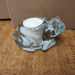 Peut inclure: Un bougeoir en forme de grenouille en verre transparent avec une bougie blanche à l'intérieur. La grenouille présente des détails et repose sur une surface en bois. Le bougeoir est un objet décoratif.
