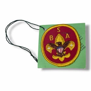 May include: A circular embroidered patch with a red background and a yellow border, featuring a gold eagle emblem with a red, white, and blue shield. The letters S, B, and A are stitched in gold. A black string is attached to a light green card.