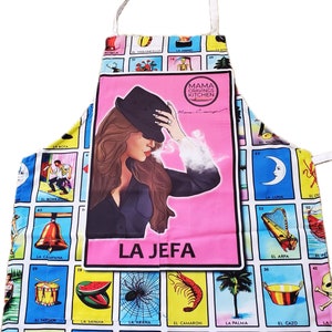 May include: A colorful kitchen apron with a white strap. The apron features a pink central panel with the words "LA JEFA" and an illustration of a woman in a hat. The apron's background is patterned with various images and text.