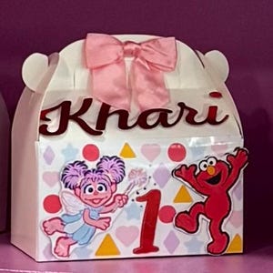 May include: A white favor box with a pink bow on top. The box is decorated with a pink, yellow, and white pattern and features a Sesame Street character, Elmo, and a character from the show, Abby Cadabby. The box is personalized with the name "Khari" and the number "1".