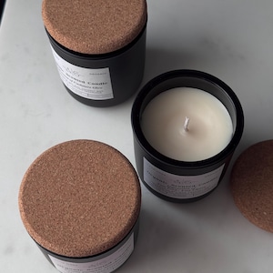 May include: Three black organic scented candles with cork lids. One candle is open, revealing a white wax interior and a wick. The labels read "Organic Scented Candle" and "Cashmere Glow".