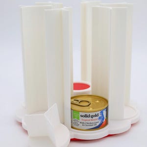May include: A white and red cat food feeder with a gold can of cat food. The feeder has a circular base with a red rim and white, vertical supports. The cat food can is labeled "solid gold" and "Tropical Blendz".