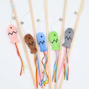 May include: A collection of six cat wand toys. Each wand has a wooden handle, a small bell, and a plush fish in various colors: pink, brown, blue, green, and gray. The fish have a zig-zag design and colorful ribbons.