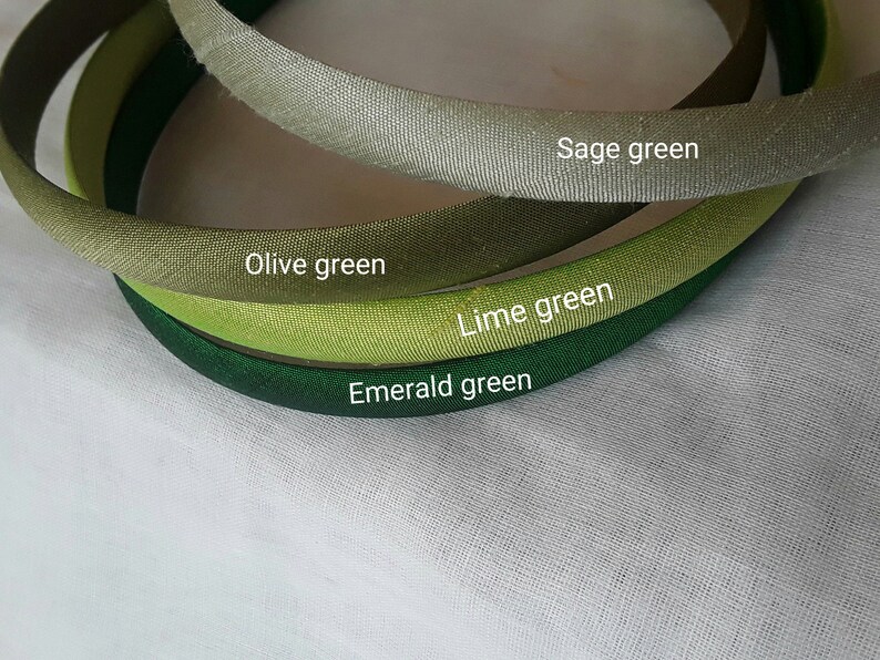 May include: Four different shades of green fabric headbands. The headbands are arranged in a circle, with the lightest shade of green at the top and the darkest shade of green at the bottom. The shades of green are sage green, olive green, lime green, and emerald green.