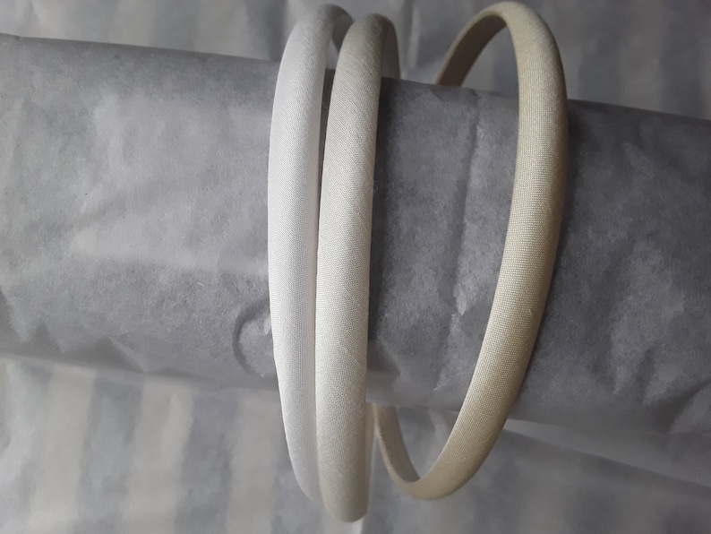 May include: Three white satin headbands, two are white and one is a slightly off-white color. The headbands are rolled up and placed on a gray and white striped surface.