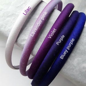 May include: Five satin headbands in shades of purple, lavender, and blue. The headbands are arranged in a gradient from light to dark, with the lightest headband labeled "Lilac", the next "Lavender Pink", then "Violet", "Purple", and finally "Bluey Purple".