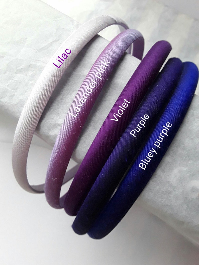 May include: Five satin headbands in shades of purple, lavender, and blue. The headbands are arranged in a gradient from light to dark, with the lightest headband labeled "Lilac", the next "Lavender Pink", then "Violet", "Purple", and finally "Bluey Purple".