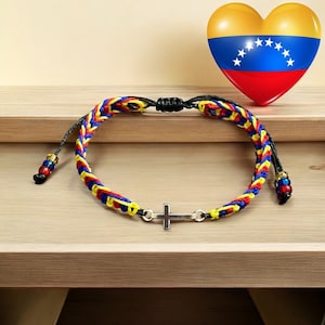 Puede incluir: Pulsera trenzada en rojo, amarillo y azul, con un dije de cruz plateada. La pulsera es ajustable con cordones negros y cuentas decorativas. Un gráfico en forma de corazón de la bandera venezolana está en el fondo.