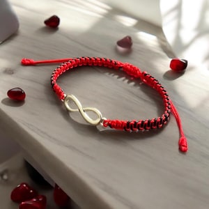 Puede incluir: Una pulsera trenzada roja y negra con un símbolo de infinito dorado. La pulsera está hecha de hilos tejidos y se muestra sobre una superficie gris claro. La pulsera es un accesorio de moda.