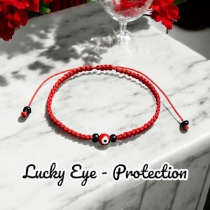 Puede incluir: Pulsera de cuerda roja con un abalorio de ojo turco negro y rojo. La pulsera está atada con un nudo y tiene una pequeña cuenta negra en cada extremo. El texto "Lucky Eye - Protection" está escrito debajo de la pulsera.