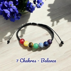 Puede incluir: Un brazalete de cuerda negra con siete cuentas de colores que representan los siete chakras. El texto "7 Chakras - Balance" está impreso debajo del brazalete.