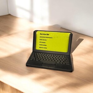 A 3d printed business card holder in the design of a laptop computer, holding a green business card, sitting on a wooden desk.