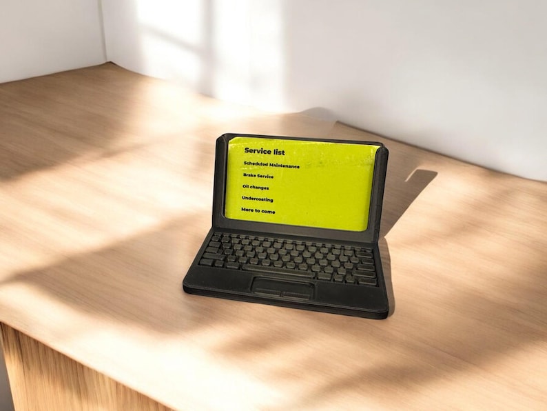 A 3d printed business card holder in the design of a laptop computer, holding a green business card, sitting on a wooden desk.