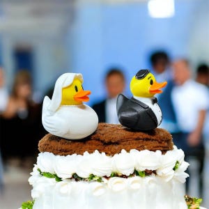 May include: A wedding cake topped with two rubber duck figurines. One duck is dressed as a bride in a white veil, and the other is a groom in a black tuxedo. The cake has white frosting and decorative flowers.