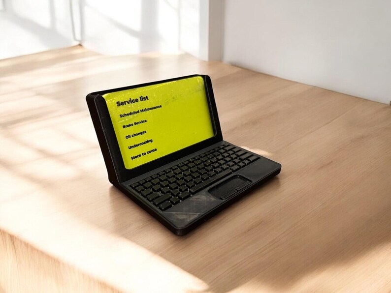 A 3d printed business card holder in the design of a laptop computer, holding a green business card, sitting on a wooden desk.