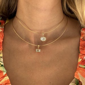 Può includere: Due delicate collane d'oro con pendenti di pietre preziose. La collana superiore presenta un pendente ovale, mentre quella inferiore ha un pendente rettangolare. Le collane sono indossate vicino al collo, creando un effetto a strati. Le pietre preziose brillano.