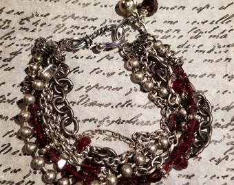 May include: A multi-strand bracelet featuring silver chains and dark red beads. The bracelet has a silver clasp.