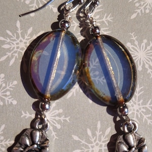 May include: A pair of dangle earrings featuring iridescent blue oval beads with silver-tone accents. Each earring has a decorative silver-tone charm with a floral design, suspended from a silver-tone hook.