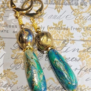 May include: Gold-tone leverback earrings with a dangling design. The earrings feature a gold bead, a blue and green marbled bead, and gold accents. The marbled beads have gold glitter accents. The background is a vintage-style paper.