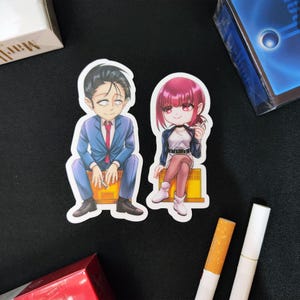 Puede incluir: Dos pegatinas de vinilo troqueladas con personajes de estilo anime. Un personaje de pelo oscuro viste un traje azul y está sentado sobre una caja amarilla. El otro tiene el pelo rosa, viste una chaqueta oscura y falda, sentado sobre una caja amarilla.