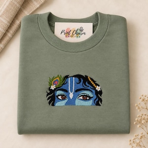 May include: An olive green crewneck sweatshirt featuring an embroidered graphic of blue eyes and dark hair with a peacock feather and white flowers. A white tag inside the collar displays the text Print Charm CREATION with a hamsa hand icon.