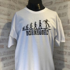 Peut inclure: T-shirt blanc avec un motif noir représentant l'évolution de l'homme, se terminant par un skateur. Le mot "EVOLUTION" est imprimé en majuscules noires. Le t-shirt a un col rond et des manches courtes.