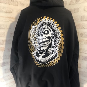 Peut inclure: Sweat-shirt à capuche noir avec un imprimé graphique or et blanc d'un crâne portant une coiffe et tenant une platine.