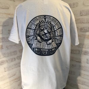 Peut inclure: T-shirt blanc avec un imprimé graphique noir et blanc d'une figure stylisée portant une coiffe et utilisant un synthétiseur. La figure est entourée d'un cercle de boutons et de molettes de synthétiseur.