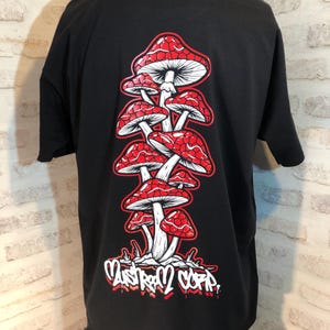 Peut inclure: T-shirt noir avec un motif de champignons rouges et blancs empilés les uns sur les autres. Le texte "Mushroom Corp" est imprimé sous les champignons.