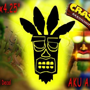 May include: A black and yellow vinyl decal featuring the Aku Aku mask from the Crash Bandicoot video game series. The mask has a tribal design with a wide mouth and a single tooth on each side. The decal measures 6 inches by 4.25 inches.