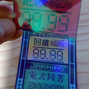 May include: A blue and green digital device with a holographic sticker on top. The sticker has the text "ALERT" and "電波障害" in Japanese. The device displays the text "回線" and "88.88" in yellow.