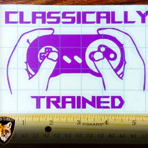 May include: A purple vinyl sticker with the text "CLASSICALLY TRAINED" and a purple silhouette of a video game controller with hands holding it.