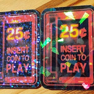 May include: Two rectangular, glittery stickers with a retro arcade game theme. Each sticker features a red border, a black background, and the text "25¢ INSERT COIN TO PLAY" in yellow. The stickers have a holographic effect.