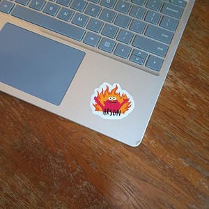 May include: A die-cut sticker featuring a red character with wide eyes and raised arms surrounded by stylized orange and yellow flames, with the word ARSON printed in black capital letters at the bottom, adhered to the palm rest of a silver laptop.