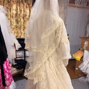 May include: A vintage ivory wedding dress with a long veil. The dress features intricate lace detailing and a high neckline. The veil is layered and extends down the back. The dress is displayed in a bridal shop.