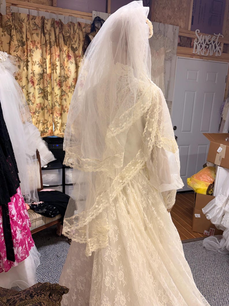 May include: A vintage ivory wedding dress with a long veil. The dress features intricate lace detailing and a high neckline. The veil is layered and extends down the back. The dress is displayed in a bridal shop.