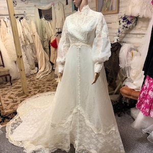 May include: A vintage white wedding dress on a mannequin. The dress features a high neckline, long sheer sleeves, and intricate lace detailing on the bodice and hem. A long train extends from the back of the dress. The dress is displayed in a vintage shop.