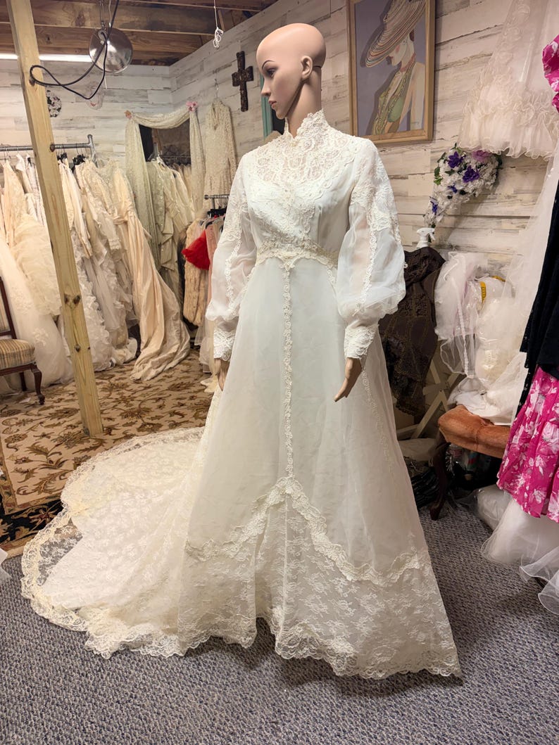 May include: A vintage white wedding dress on a mannequin. The dress features a high neckline, long sheer sleeves, and intricate lace detailing on the bodice and hem. A long train extends from the back of the dress. The dress is displayed in a vintage shop.