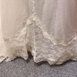May include: Ivory-colored wedding dress with a lace hem. The dress features a layered design with a sheer overlay and intricate lace detailing along the bottom edge. The lace trim has a scalloped edge.